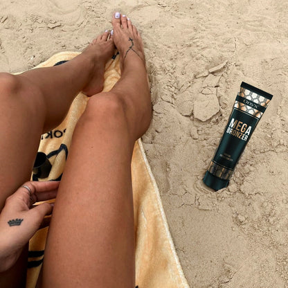 Eine Frau mit gebräunten Beinen sitzt am Strand auf einem Handtuch, neben ihr liegt eine Tube Onyx Mega Bronzer