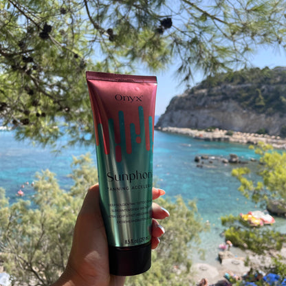 Eine Hand hält eine Tube Onyx Sunphoria Bräunungsbeschleuniger vor einer sonnigen Meereslandschaft mit klarem blauen Wasser und grünen Bäumen