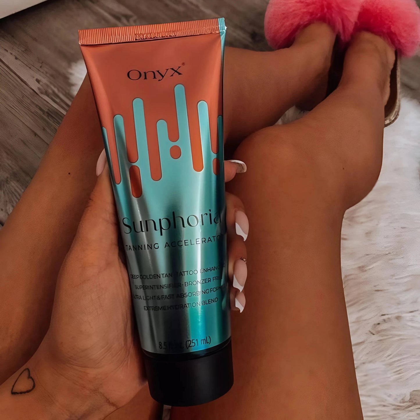 Eine gebräunte Person sitzt auf einem weißen Fellteppich, trägt rosafarbene Hausschuhe und hält eine Tube Onyx Sunphoria Bräunungsbeschleuniger in der Hand