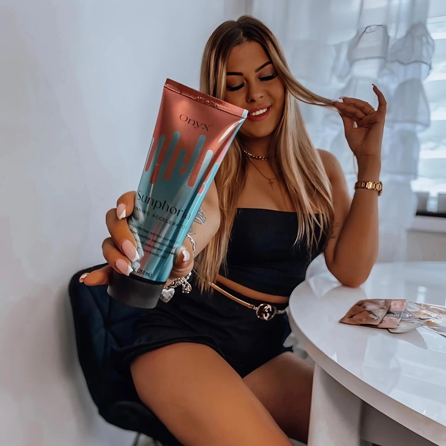 Eine lächelnde blonde Frau sitzt an einem weißen Tisch, trägt ein schwarzes Outfit und hält eine Tube Onyx Sunphoria Bräunungsbeschleuniger in die Kamera