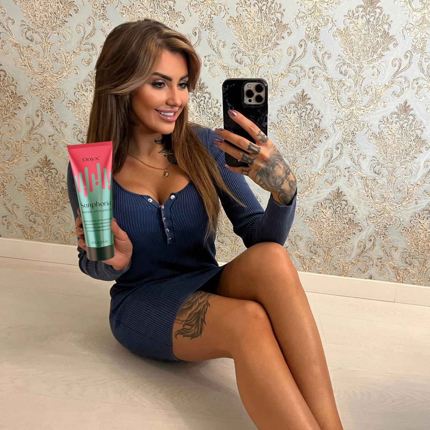 Eine Frau mit langen Haaren und Tattoos sitzt auf dem Boden, trägt ein blaues Kleid und hält eine Tube Onyx Sunphoria Bräunungsbeschleuniger, während sie ein Selfie macht