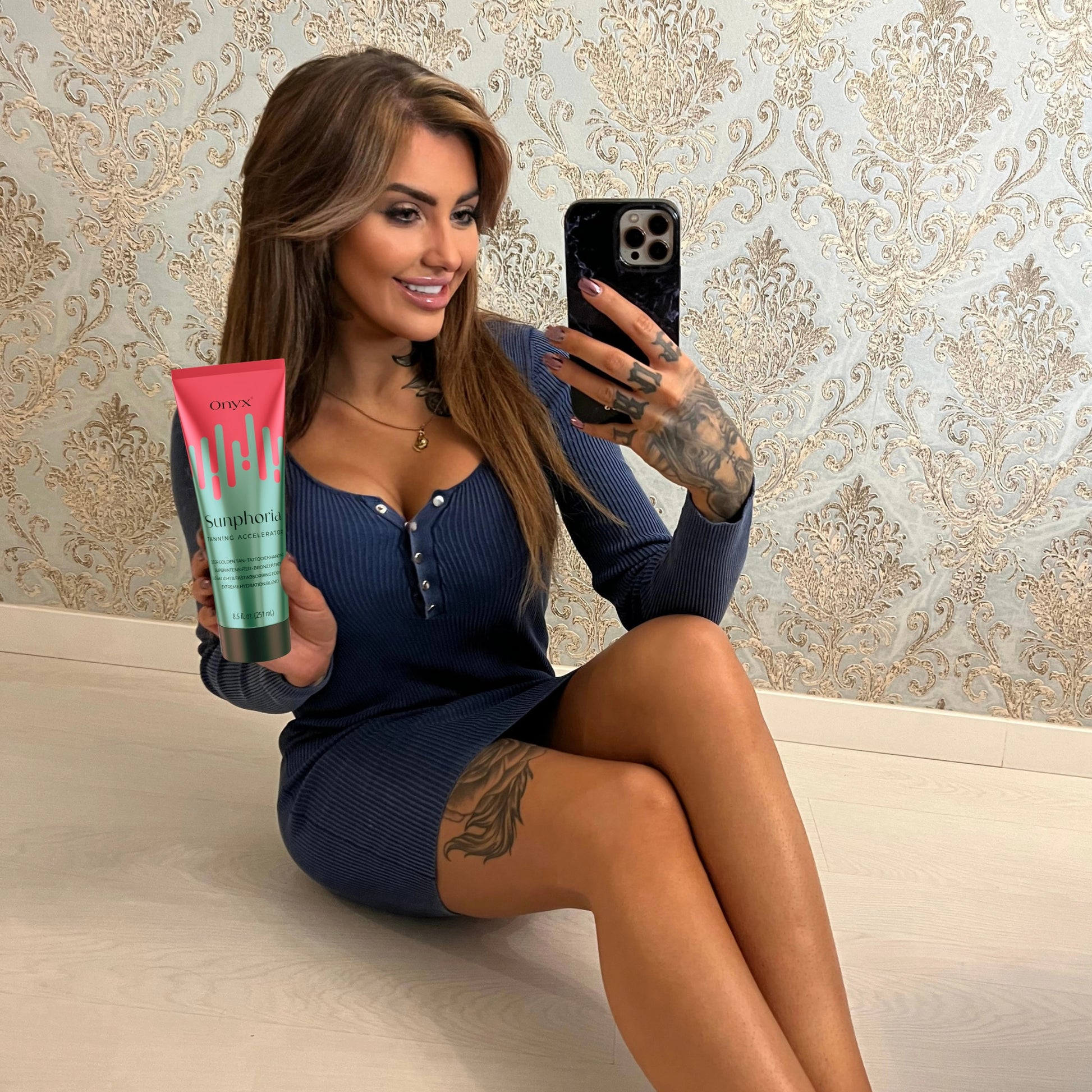 Eine Frau mit langen Haaren und Tattoos sitzt auf dem Boden, trägt ein blaues Kleid und hält eine Tube Onyx Sunphoria Bräunungsbeschleuniger, während sie ein Selfie macht