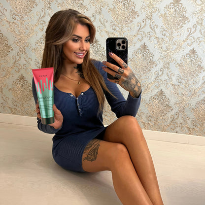 Eine Frau mit langen Haaren und Tattoos sitzt auf dem Boden, trägt ein blaues Kleid und hält eine Tube Onyx Sunphoria Bräunungsbeschleuniger, während sie ein Selfie macht