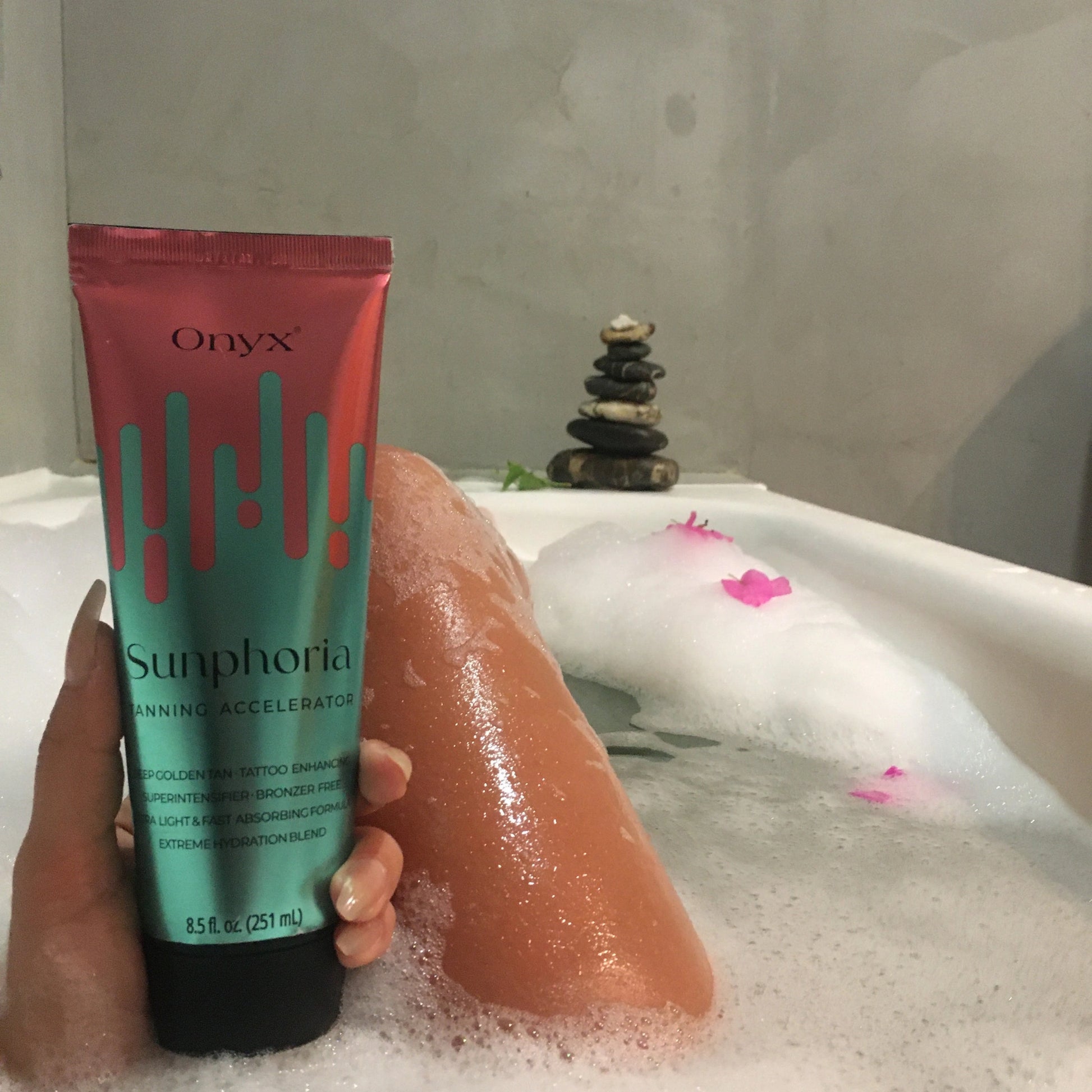 Eine Person liegt in einer Badewanne mit Schaum und hält eine Tube Onyx Sunphoria Bräunungsbeschleuniger; im Hintergrund liegen kleine Steine und Blütenblätter